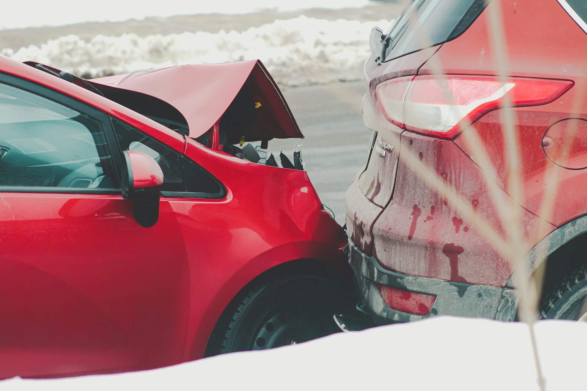 A low-angle, close-up shot of a rear-end collision between two red cars on a snowy or icy road. The car in the foreground has severe damage to its front end, with the hood crumpled and pushed upwards. The car in the background has minor damage to its rear bumper. The road and surrounding area are covered in a thin layer of snow.