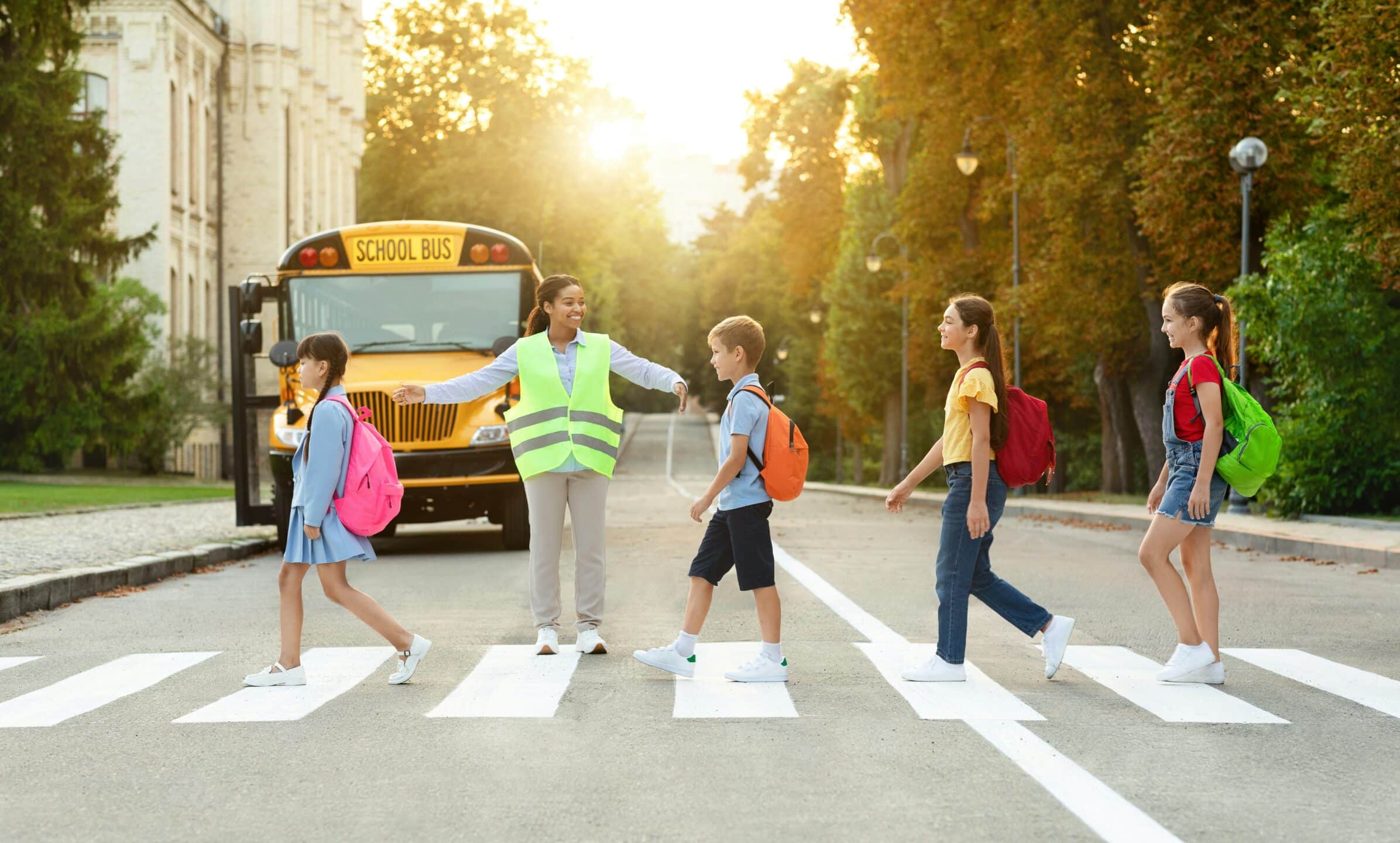 A school crossing guard wearing a safety vest is stopping traffic, represented by a yellow school bus in the background, to allow four elementary school children with backpacks to cross the street at a marked crosswalk. This image illustrates the need for caution in school zones to prevent school zone accidents and crosswalk collisions.