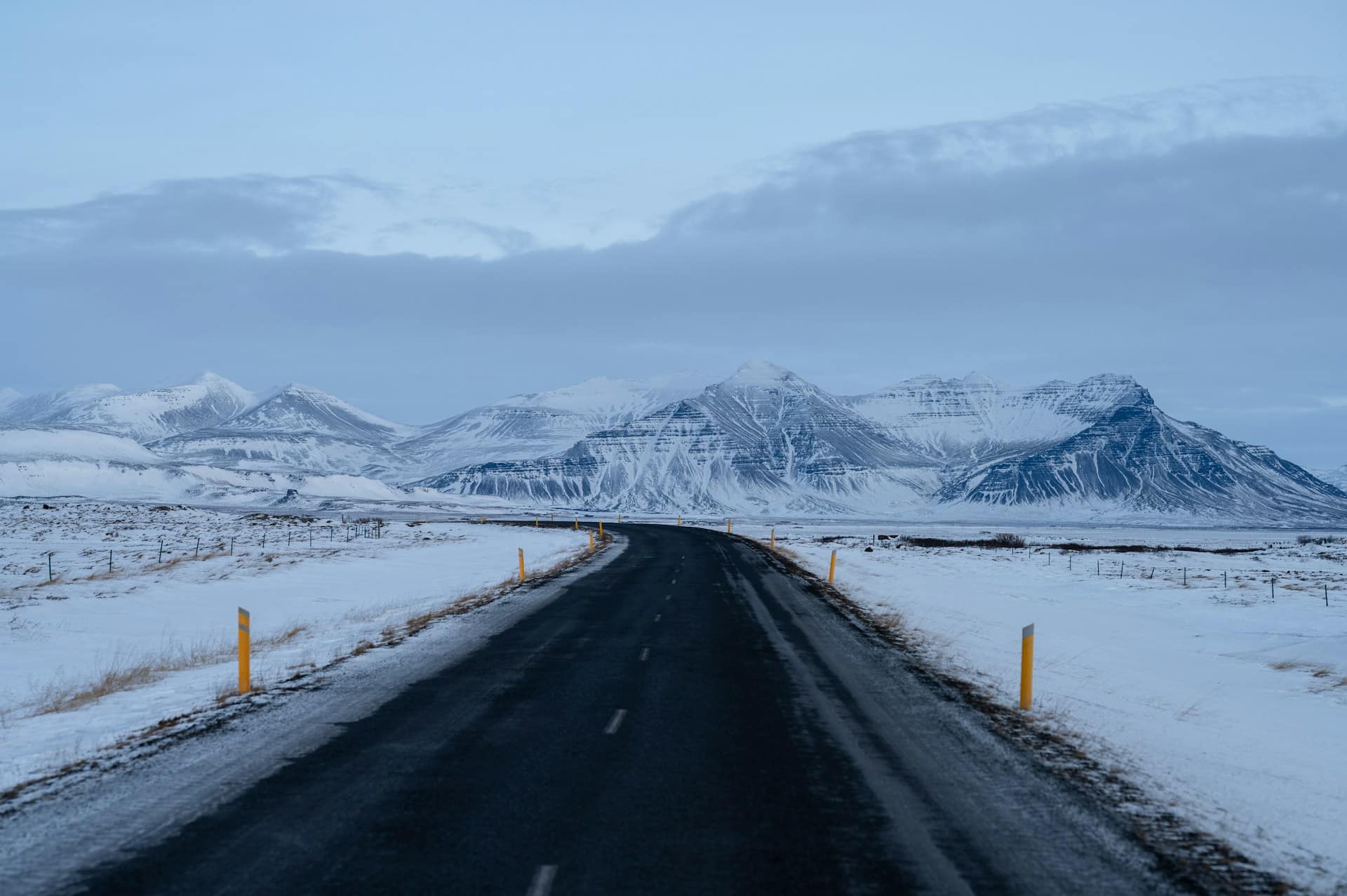 A black stretch of road splits the snow-covered ground and curves toward snowy mountains under a blue sky with a distinct line of grey cloud rising up from behind the mountains, representing winter car accidents.