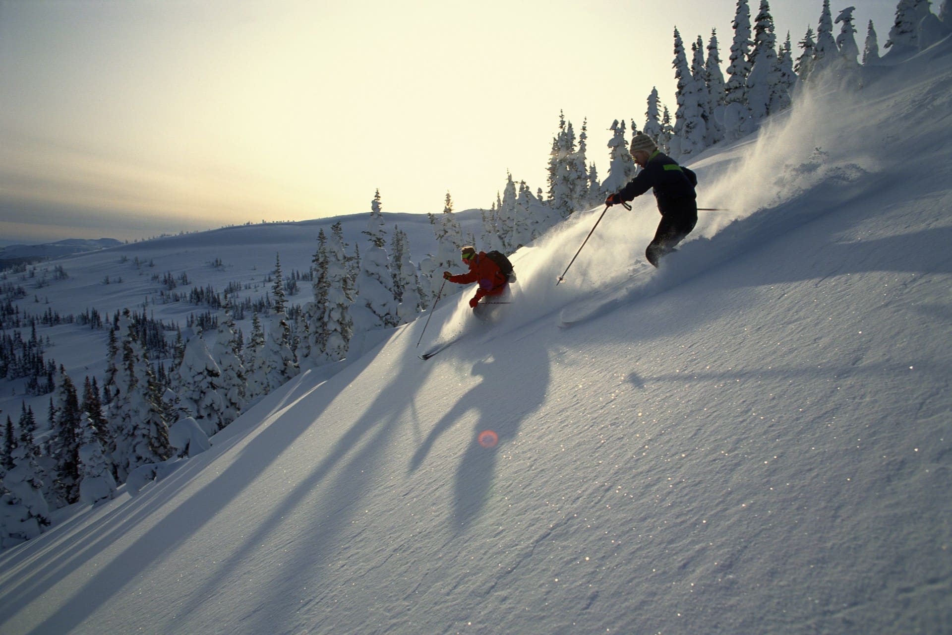 Two skiers sail down a steep snow-covered hill, sending up puffs of snow in their wake, while the sunsets behind them and over snow-covered fir trees and hills in the background, representing ski hill injuries.