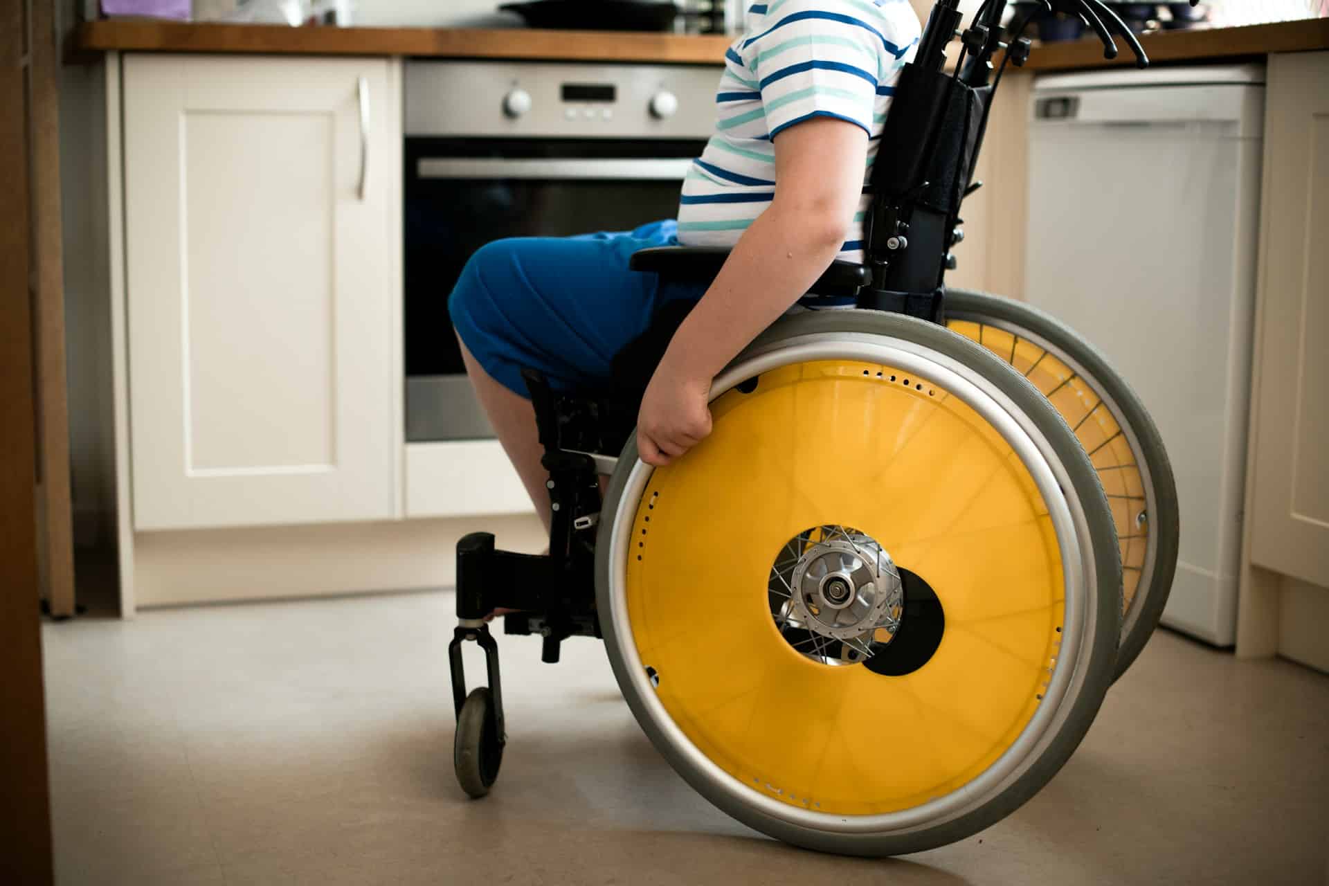 A child is shown from the shoulders down in a wheelchair with yellow covers over the wheels' spokes in a kitchen, representing the different types of cerebral palsy and their impact on medical malpractice claims.
