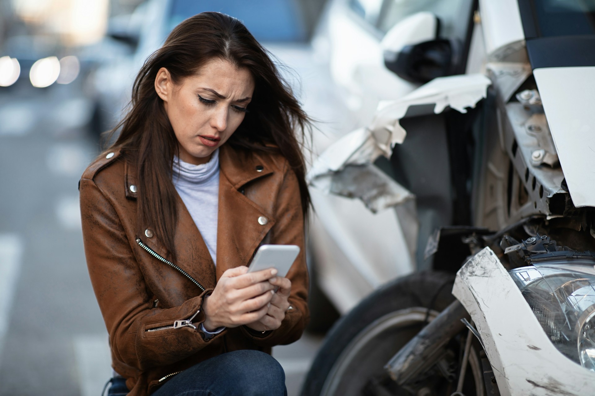 A woman kneels next to her damaged car after a collision and makes a call on her cell phone, representing broker liability for denied insurance claims.