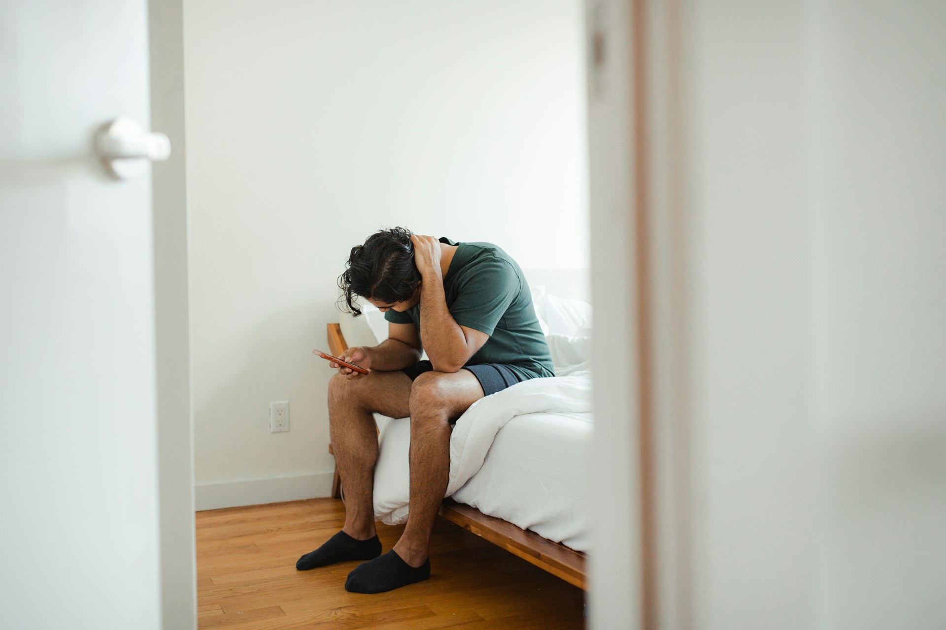 A man with dark hair wearing shorts and holding a phone, sitting at the side of his bed, visibily stressed, represeting PTSD for people in Alberta after an accident.