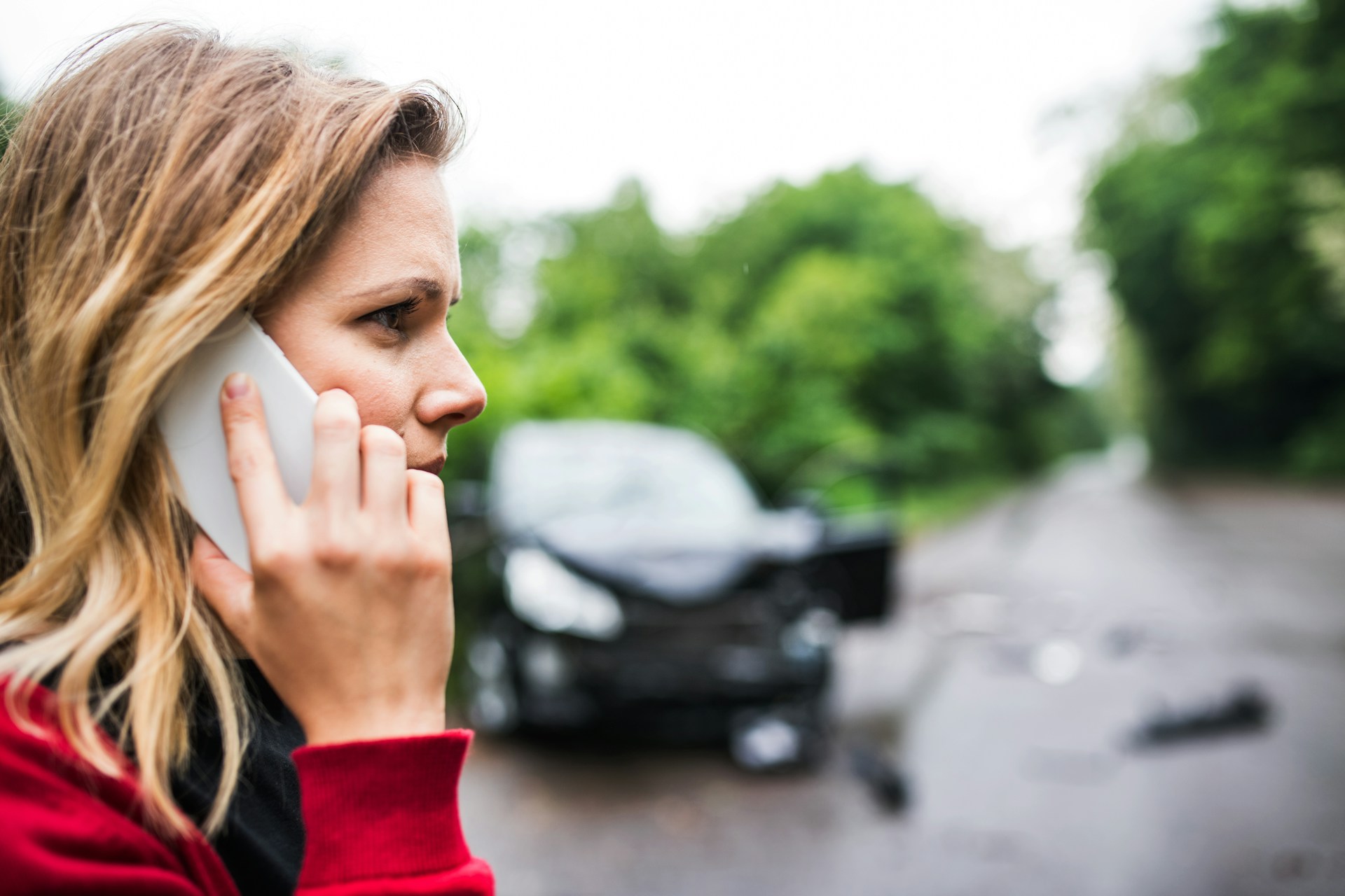 A close-up of a woman's profile with her phone to her ear and a blurred car damaged from a collision in the background, representing out-of-town car accidents in Alberta.