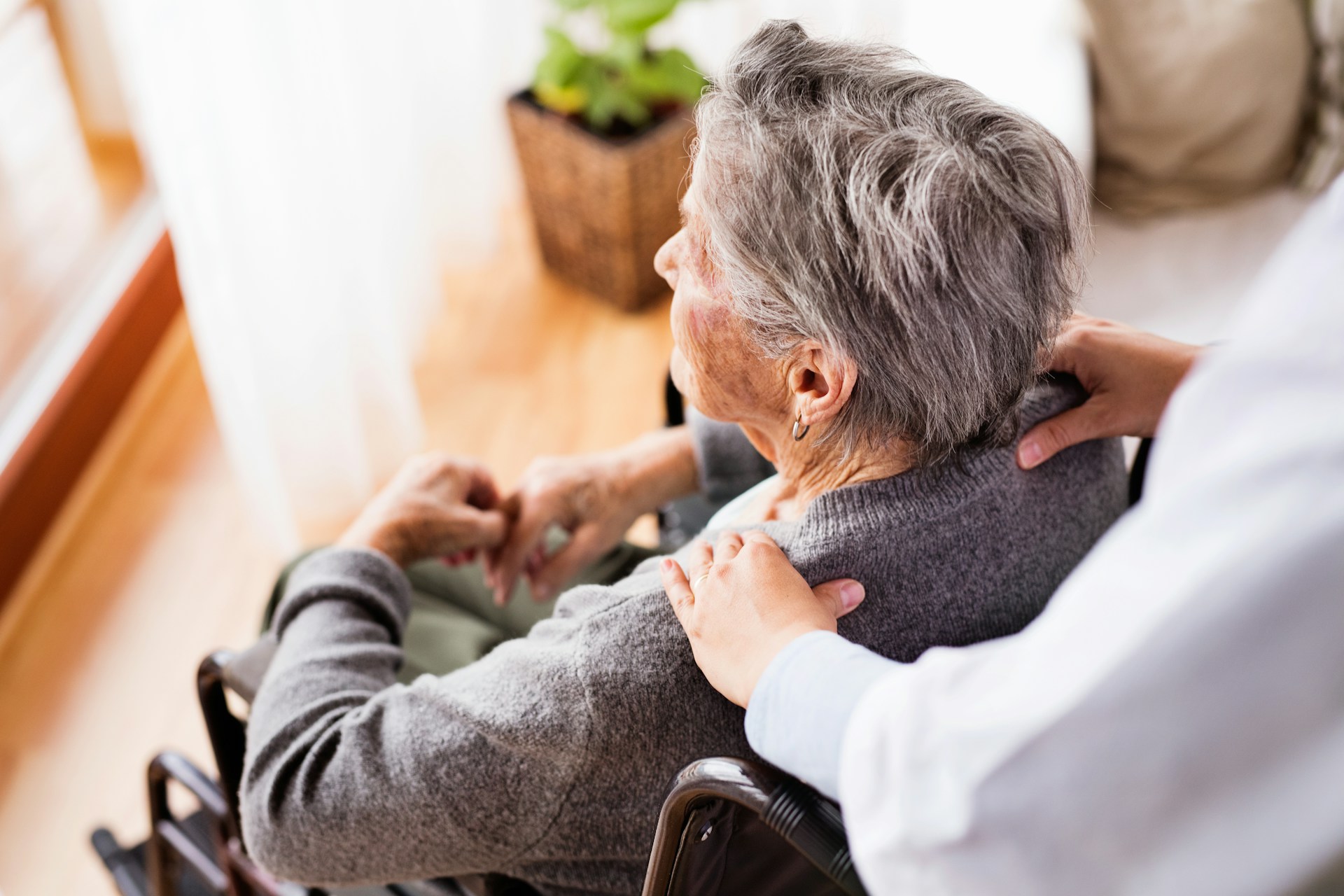 A woman in a wheelchair gazes out a nursing home window while someone stands behind her gripping her shoulders, representing resident-on-resident violence in Alberta nursing homes.