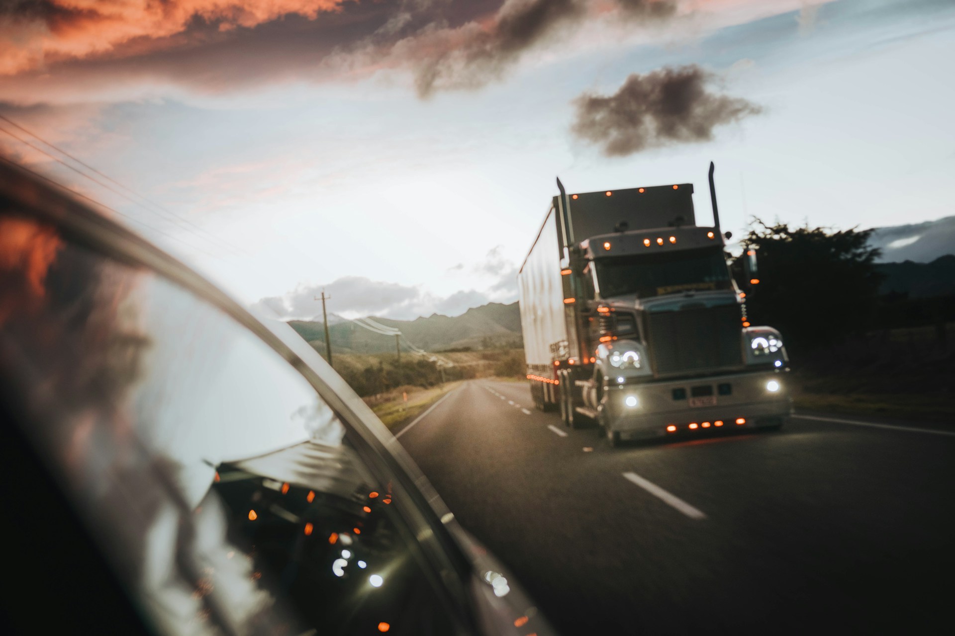 A transport truck travels down a highwy at dusk with the rear corner of the car in front of it visible, representing truck driver fatigue.